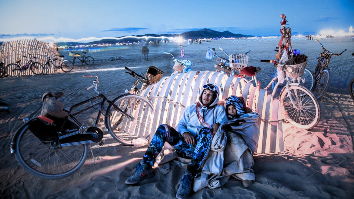 Two burners bundled up in warm jackets resting against a bicycle rack at dusk on the playa