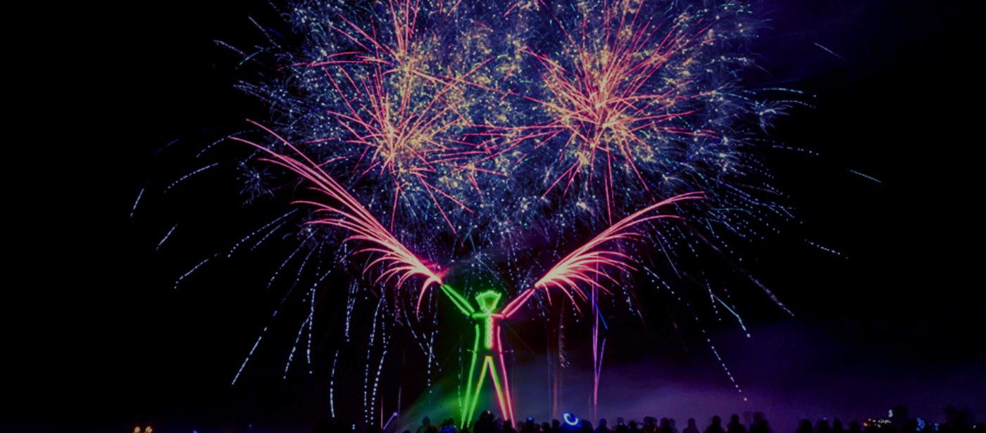 Fireworks bursting over a glowing green and red humanoid light figure with arms raised, silhouettes of the crowd below