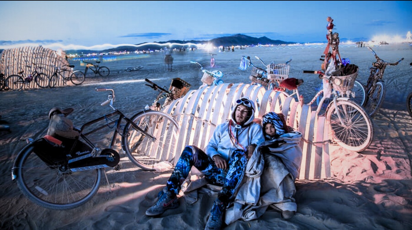 Two people bundled in blankets at dusk beside a lit wooden bike shelter